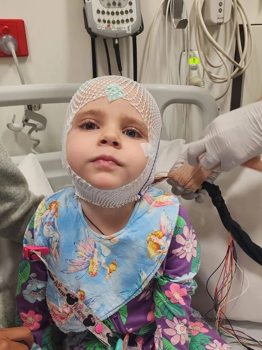 A toddler with EEG electrodes on her head, sitting up in a hospital bed during testing.