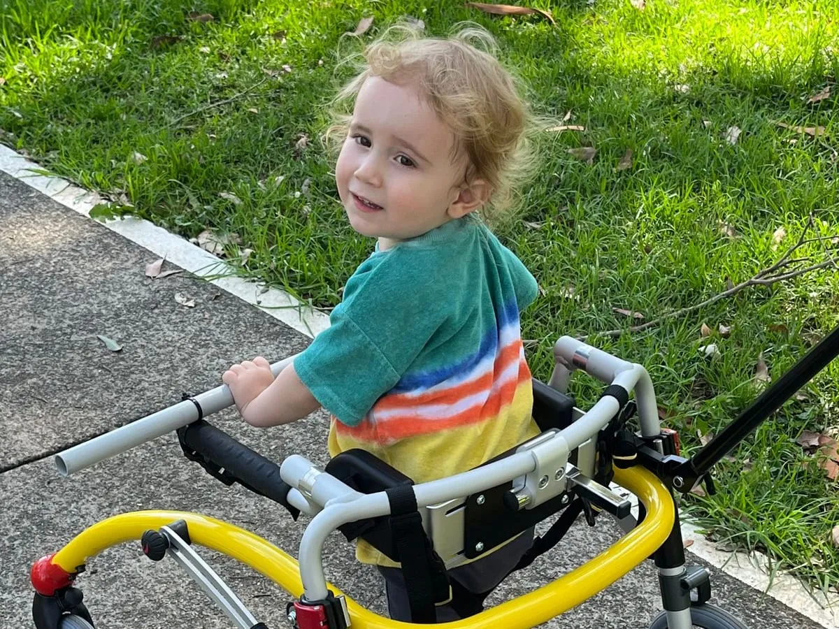 A young child with an STXBP1 disorder smiling over their shoulder while walking in a supported walker.