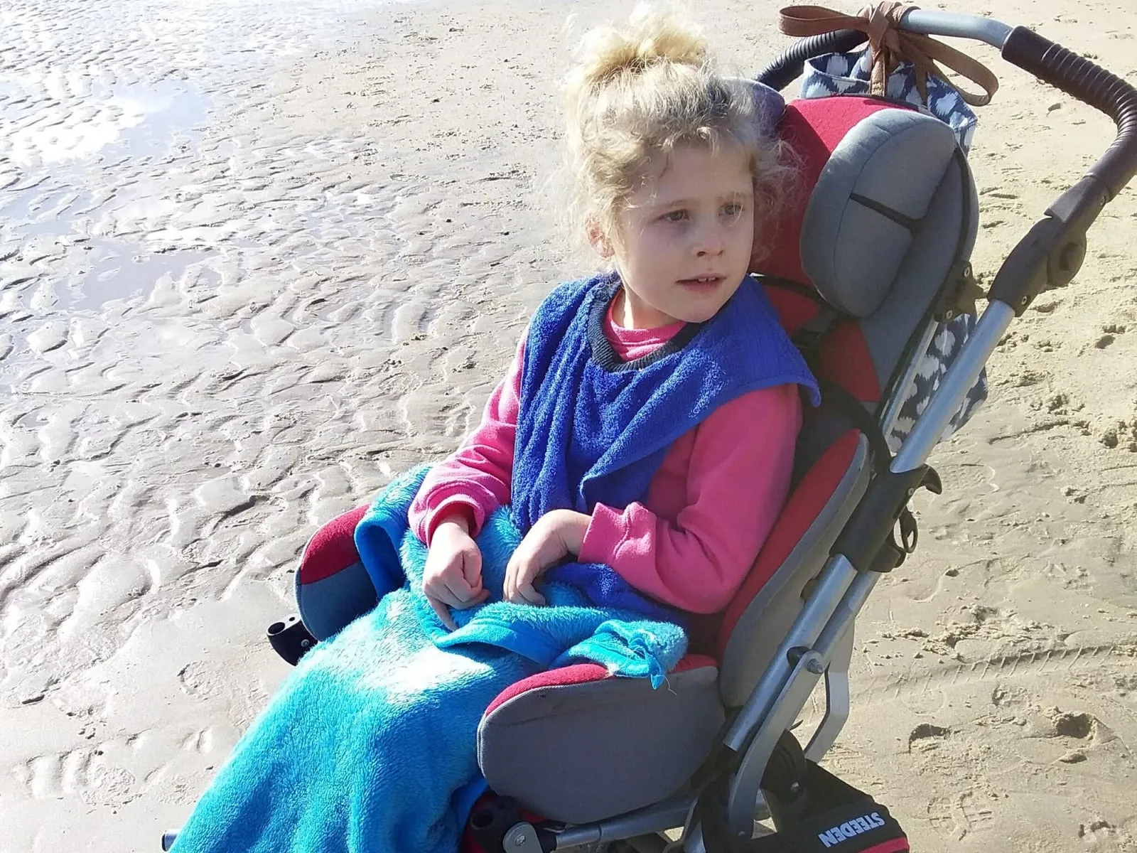 Annabelle sitting in her supportive wheelchair on the beach, wrapped in a towel.