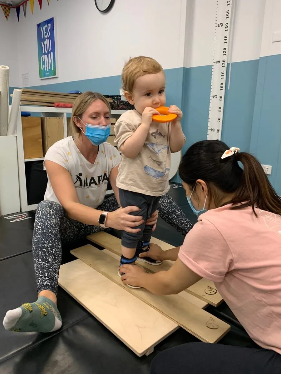 A toddler stepping onto a balance board, supported by two therapists.