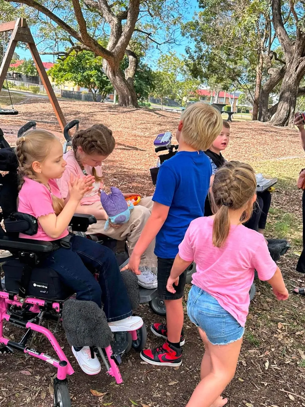Children from STXBP1 Australia families, including a child in a pink wheelchair, gathered together at a park.