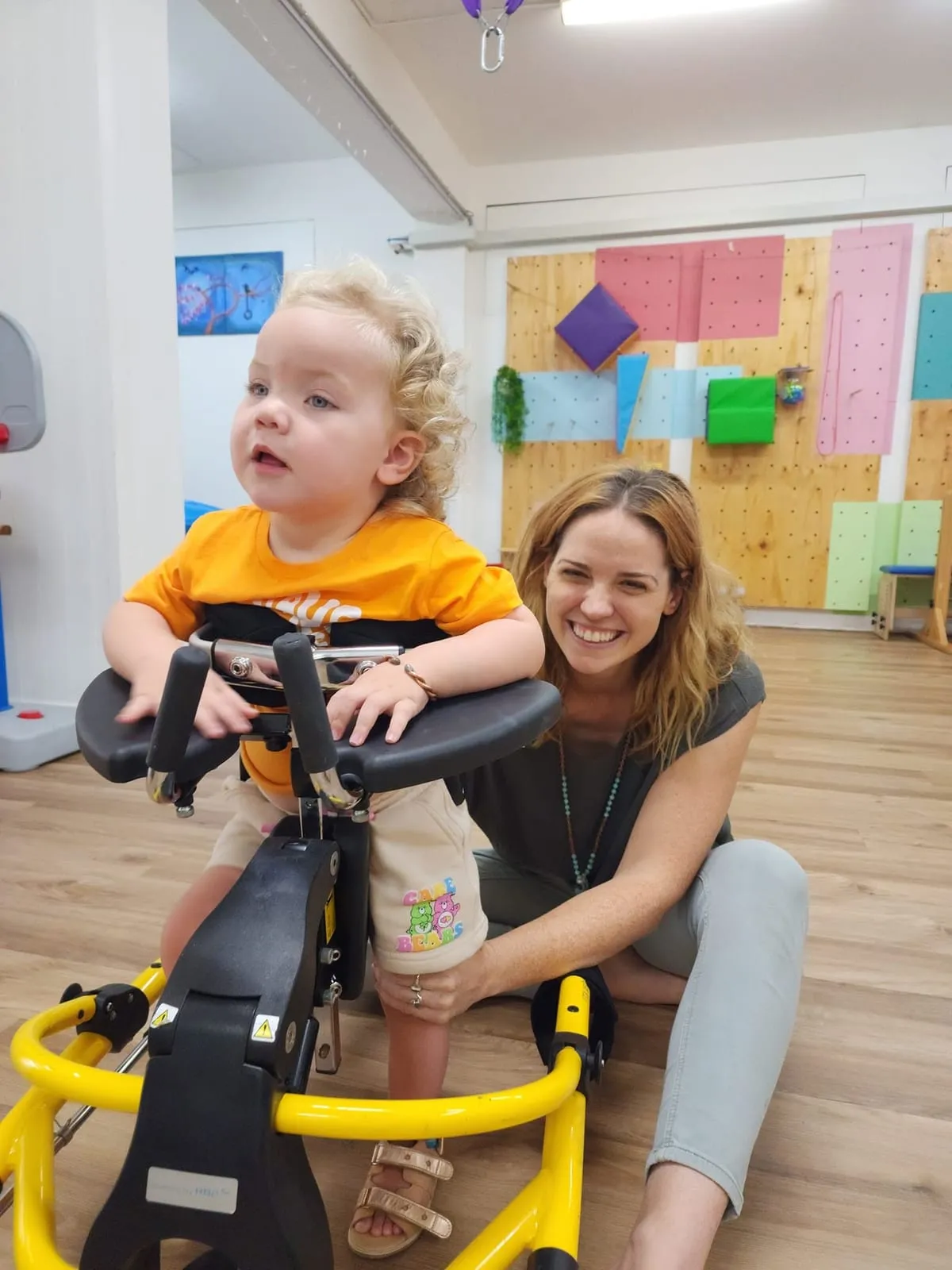 A smiling mum sitting beside her toddler, who is standing in a supported walker.