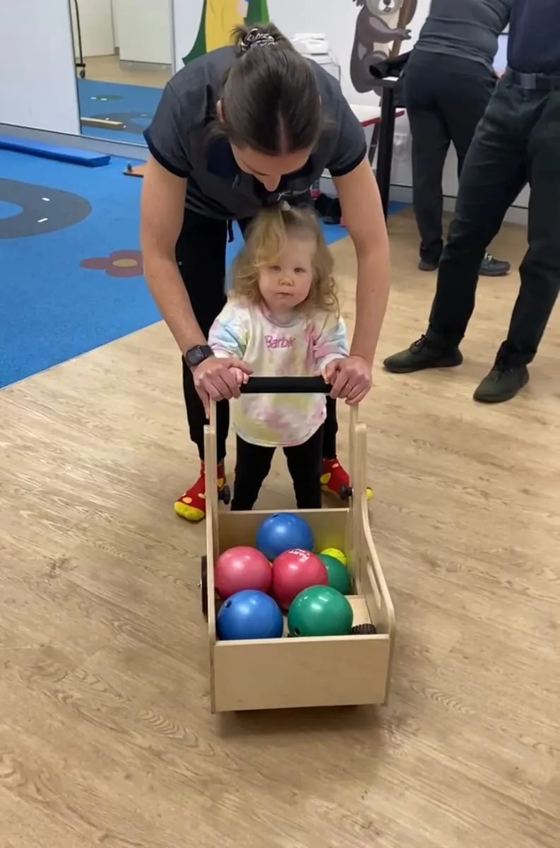 A little girl walking behind a push cart, steadied by her therapist.